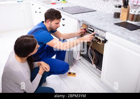 Woman Looking At Male Technician Checking Dishwasher With Digital Multimeter In Kitchen Stock Photo