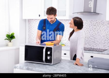 Pretty Woman Standing Near Repairman Repairing Oven Using Digital Multimeter In Kitchen Stock Photo