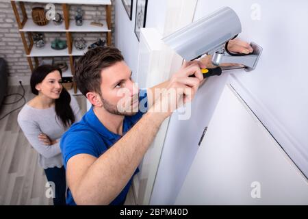 Smiling Woman Looking At Male Technician Adjusting CCTV Camera On Wall With Screwdriver Stock Photo
