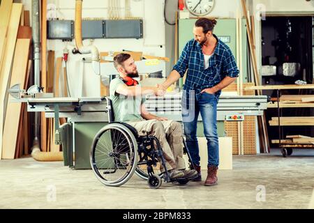 disabled worker in wheelchair in a carpenter's workshop with his colleague in conversation Stock Photo