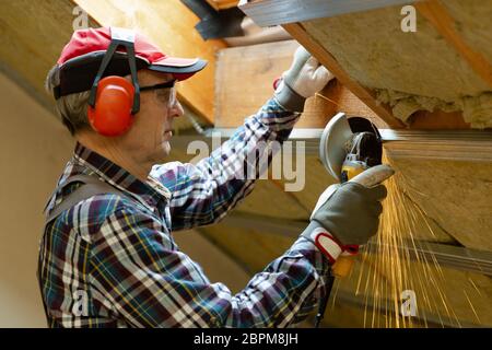 Man fixing metal frame using angle grinder on attic ceiling covered with rock wool Stock Photo