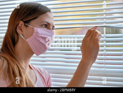 Young woman in pink home made cotton virus face mask, looking through window blinds outside. Quarantine or stay at home during coronavirus covid-19 ou Stock Photo