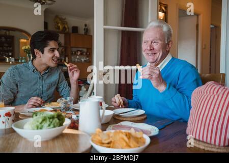 Senior man is having lunch at home with his grandson. They are eating sandwiches and potato chips. Stock Photo