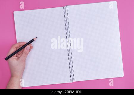 human hand holds black wooden pencil over empty open notebook, top view, pink background Stock Photo