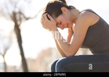 Profile of a sad teen crying desperately sitting on a bench in a park Stock Photo