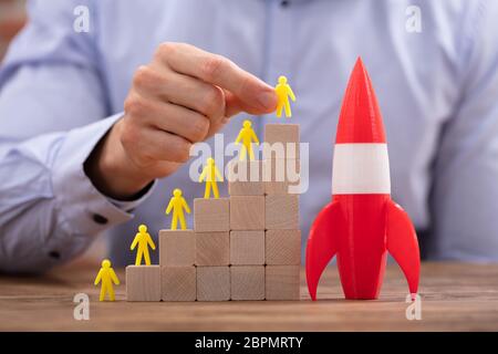 Close-up Of Person's Hand Placing Yellow Human Figure On Top Of Wooden Blocks Besides Rocket Stock Photo