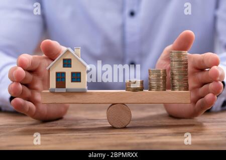 Small House Model And Stacked Golden Coins Balancing On Wooden Seesaw Stock Photo