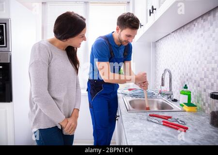 Happy Woman Looking At Male Plumber Using Plunger In The Kitchen Sink Stock Photo