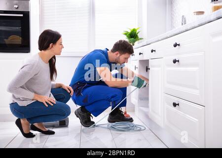 Woman Looking At Male Plumber Cleaning Clogged Sink Pipe With Drained Cable In Kitchen Stock Photo