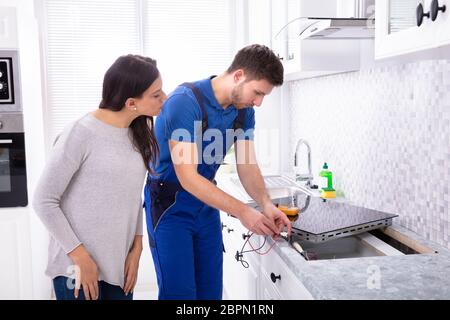 Male Repairman Installing Induction Cooker With Woman Standing In Kitchen Stock Photo