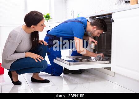 Young Woman Looking At Repairman Repairing Dishwasher In Kitchen Stock Photo