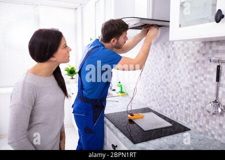 Male Repairman Checking Extractor Filter With Digital Multimeter While Woman Standing In Kitchen Stock Photo