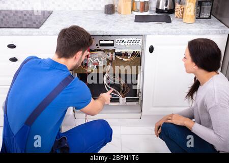 Woman Looking At Male Technician Checking Dishwasher With Digital Multimeter In Kitchen Stock Photo