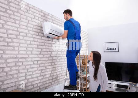 Young Woman Looking At Male Technician Fixing Air Conditioner On Brick Wall Stock Photo