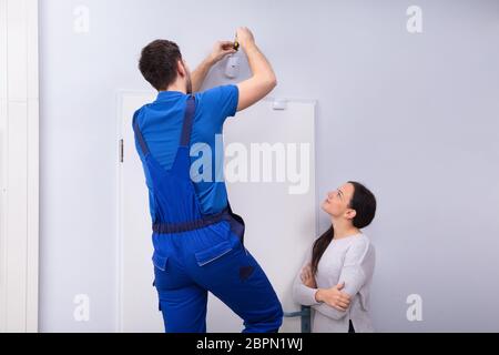 Happy Woman Looking At Electrician Installing Security System Door Sensor On Wall Stock Photo