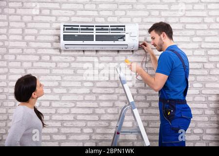 Smiling Woman Looking At Male Technician Repairing Air Conditioner With Digital Multimeter At Home Stock Photo