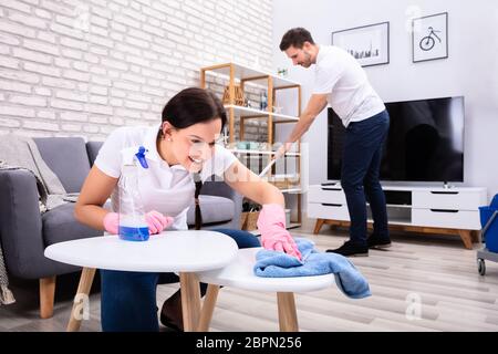 Two Smiling Young Workers Cleaning The Table And Mopping Floor In The Living Room Stock Photo