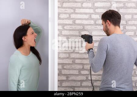 Woman Shouting Behind The Wall With Neighbor Man Using Power Drill On White Brick Wall Stock Photo