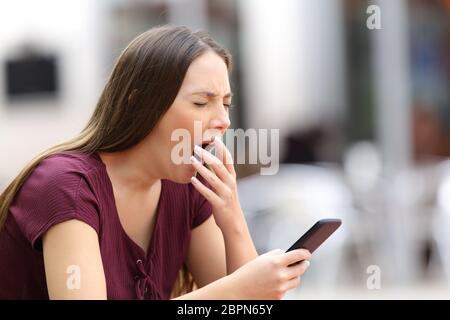 Woman yawning bored with on line content holding a mobile phone sitting on a bench in the street Stock Photo