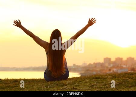 Back view backlight portrait of a single cheerful woman raising arms watching the city at sunset with a warm light in the background Stock Photo