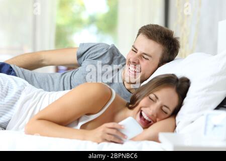 Happy couple lying on the bed laughing watching smart phone content Stock Photo