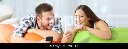 Two happy friends sharing media content on line with their smart phones lying on colorful puff in a house interior Stock Photo