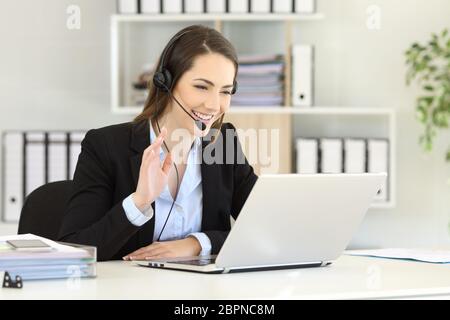 Happy telemarketer having a video conference with a laptop waving with her hand at office Stock Photo