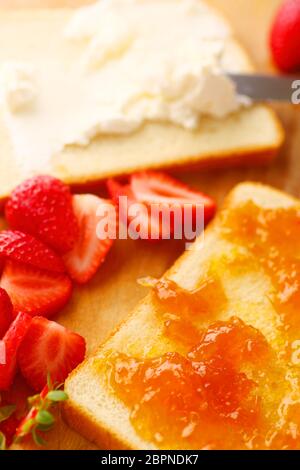 Spreading cream cheese on white bread with jam and strawberries alongside Stock Photo