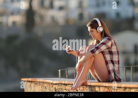 Happy teenage girl watching video on a smart phone sititng on a ledge in a town at sunset Stock Photo