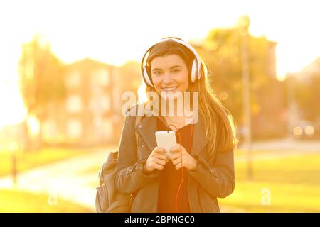 Happy teenage girl holding smart phone listening to music looking at you in a park at sunset Stock Photo