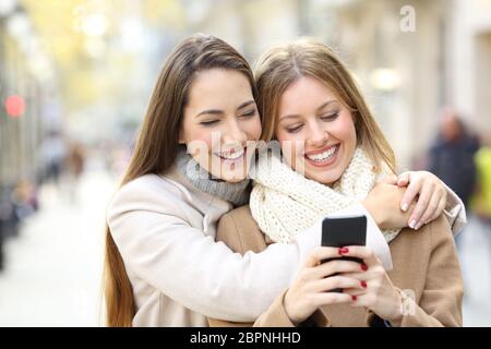 Front view portrait of two cheerful friends watching smart phone content in winter on the street Stock Photo