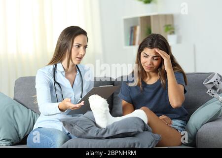 Doctor and sad patient with plaster foot sitting on a couch in the living room at home Stock Photo