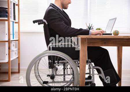 Disabled Businessman Sitting On Wheelchair Using Laptop At Workplace Stock Photo