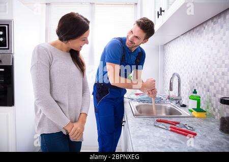 Happy Woman Looking At Male Plumber Using Plunger In The Kitchen Sink Stock Photo