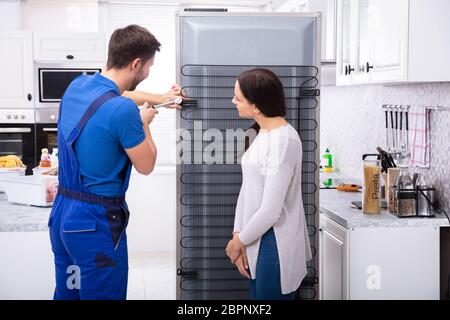 Woman Looking At Technician Repairing Refrigerator With Screwdriver In Kitchen Stock Photo