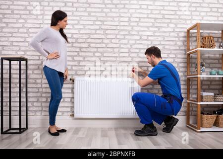 Beautiful Woman Looking At Young Male Plumber In Uniform Installing Radiator With Screwdriver Stock Photo