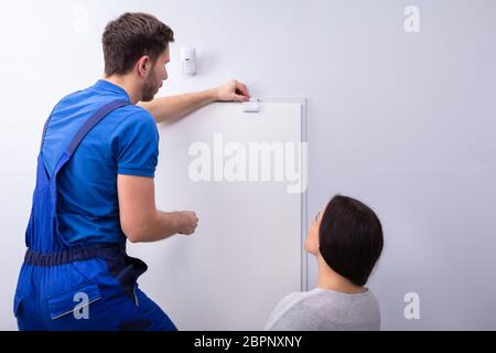 Happy Woman Looking At Electrician Installing Security System Door Sensor On Wall Stock Photo
