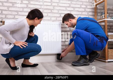 Crouching Woman With Serviceman Turning Radiator Bleed Valve To Release Air With Cup Stock Photo