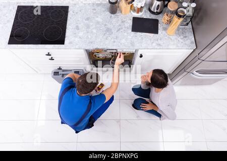 Woman Looking At Male Technician Checking Dishwasher With Digital Multimeter In Kitchen Stock Photo