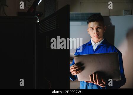 Young man is looking at the camera while in the server room with a laptop. Stock Photo