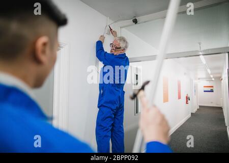 Senior electrician is talking to his trainee while fitting a security camera in the corridor of an office building. Stock Photo