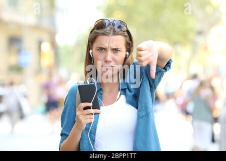 Front view portrait of an annoyed girl listening to music with thumbs down in the street Stock Photo