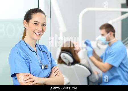 Dentist female with crossed arms wearing blue coat posing and looking at you at consultation with a doctor working with a patient in the background Stock Photo