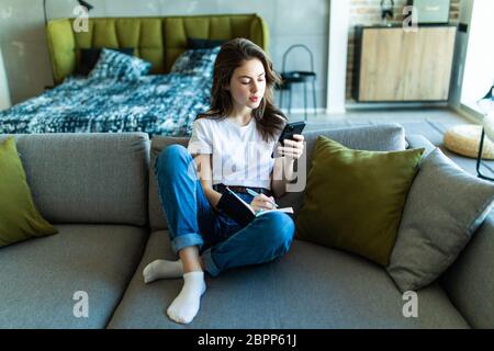 Close up of smiling woman sitting near sofa, writing in weekly planner and surfing phone. Working at home. Stock Photo