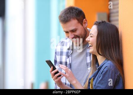 Happy couple laughing loud checking smart phone online content in a colorful street Stock Photo