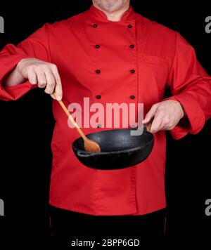 cook in red uniform holding an empty  black frying pan and wooden spoon, black background Stock Photo
