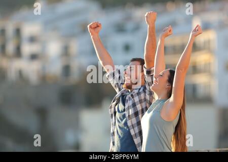 Excited couple celebrating vacation raising arms in a town at sunset Stock Photo