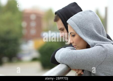 Side view of a sad couple of teens looking down after break up in a balcony of a house with an urban background Stock Photo
