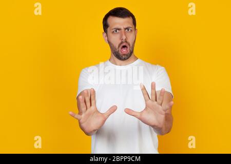 A young man with a beard in a white T-shirt standing on an isolated yellow background, a frightened man stretched out his hands in front with a stop gesture of an expression of fear screaming in shock Stock Photo