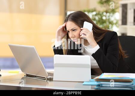 Worried executive talking on the phone trying to solve problems with multiple devices sitting in a desktop at office Stock Photo